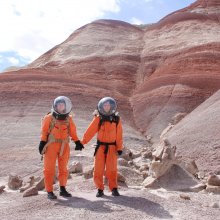 Two women in spacesuits in a barren landscape holding hands