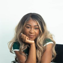 A young Black woman with long blonde hair rests her head on her chin and smiles softly into the camera.