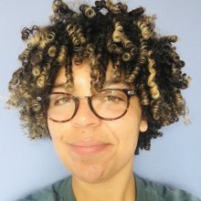 A person with curly, multi-tonal hair poses against a soft blue background, wearing a green shirt.