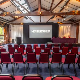 Event venue with rows of red chairs facing a stage, projection screen labeled “WATERSHED,” and panel table.