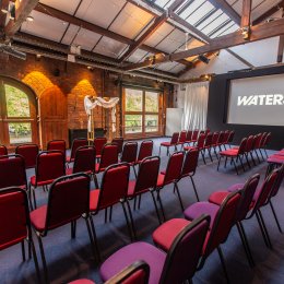 Event venue with rows of red and purple chairs, exposed brick walls, wooden beams, string lights, and a large screen reading “WATERSHED.”