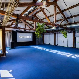 Industrial event space with exposed wooden beams, blue carpet, hanging plants, projector, and a screen displaying “WATERSHED.”