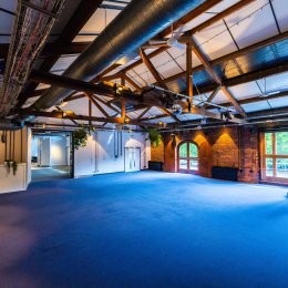 Spacious industrial-style event hall with exposed beams, ductwork, blue carpet, brick walls, windows, and stage equipment.