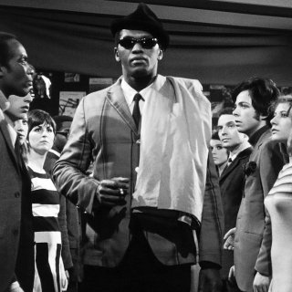 Black and white film still of a man wearing a trilby and shades as a group of people watch him walk through the room