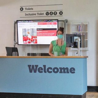 Photo of woman in the cinema foyer at Watershed, wearing a mask with a welcome sign on the reception desk in from of her