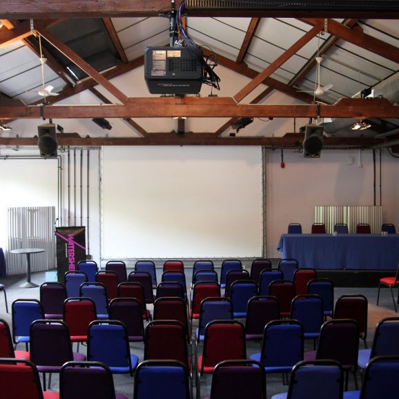 Photo of Waterside 3 at Watershed – a large room with chairs facing a projector screen, a speakers podium and top table