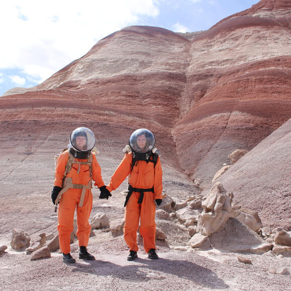 Two women in spacesuits in a barren landscape holding hands