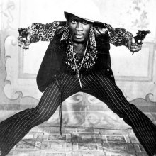 A photo of a man in seventies clothing with legs wide apart brandishing two pistols