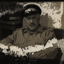 An image of a damaged piece of film reel. We can make out a man in uniform, looking seriously at the camera. The surface of the reel has come away, leaving grey grazes across the surface. 