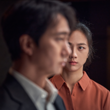 A woman stares intensely at a man, who faces the camera side-on in the foreground. 