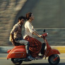 Two people ride on a red scooter, the background a blur.
