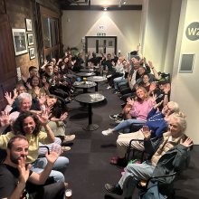 hoto of Deaf Conversations About Cinema discussion group at Watershed. A large group of people sitting in a long corridor together - waving their hands in the air after the discussion.