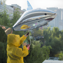Person in a yellow raincoat holding a large silver shark-shaped balloon near a river with trees and city buildings in the background.