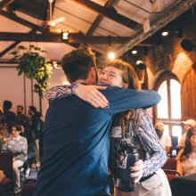Two people hug and smile in a room with exposed brick walls, while others sit and watch in the background.