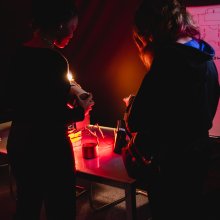 Two people examine a small object at a cluttered worktable under red light, beside a whiteboard covered in sketches and notes; the mood is focused and tense.