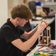 A focused young man in a black polo carefully assembles an electronics project at a workbench, surrounded by wires and tools in a busy workshop.