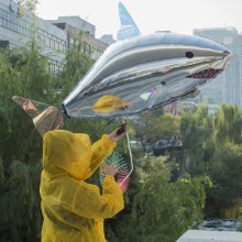 A person dressed in a yellow mac holds up a shiny silver balloon. It is a shark.