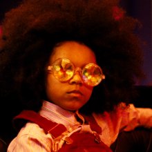 A child wears large, round mirrored sunglasses and dungarees and looks towards the camera. 