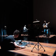 A group of musicians in a dark studio. One plays guitar, one drums and a piano in the background.