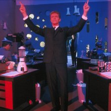 Daivd Bowie pointing his hands in the air standing amongst desks.