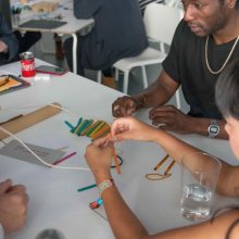 A diverse group if people are doing an activity together on a white tables with string and colourful lollipop sticks.