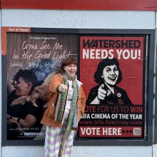 An outdoor poster site - a person stands to the side smiling and pointing in the same pose as the person in the poster which also has the words' Watershed Nees