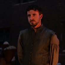 A young man stands in dim light, wearing a historical tunic with an embroidered collar, while figures gather in the background.