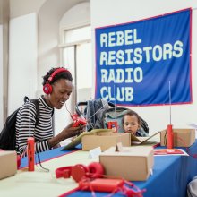 A young Black women sits smiling with red headphones on and a banner above her reads Rebel Resistors Radio Club. 