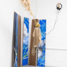 A minimalistic setup features headphones on a wall mount, a framed artwork above a yellow cloth-draped shelf, and two brown vases with pampas grass below.