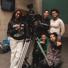Five young people posing with a professional ARRI camera on a tripod in an indoor setting with concrete walls.