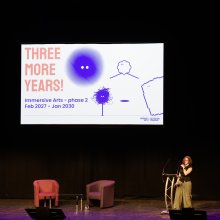 Speaker stands on a dimly lit stage beside a slide reading “THREE MORE YEARS!” with colorful drawings, creating an optimistic, creative tone.