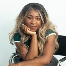 A young Black woman with long blonde hair rests her head on her chin and smiles softly into the camera.
