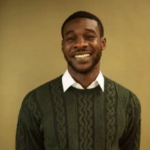 A person wearing a green cable-knit sweater over a white collared shirt stands against a neutral background.