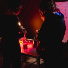 Two people examine a small object at a cluttered worktable under red light, beside a whiteboard covered in sketches and notes; the mood is focused and tense.