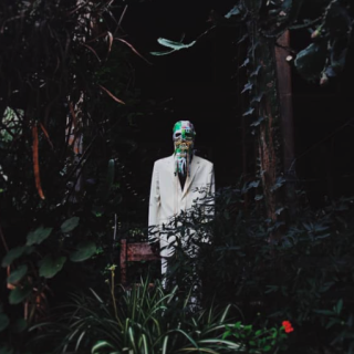 Still of a person in as white suit, wearing a mask standing in a rich green plant setting