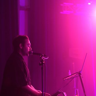 A performer sings into a microphone on stage, bathed in vibrant pink and purple lighting, creating a dramatic and immersive atmosphere.