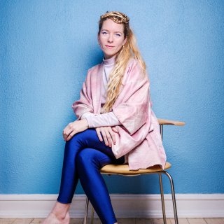 A woman with blonde hair sits cross legged on a char, looking at the camera.