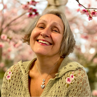 A portrait head shot photograph of Michelle Rumney, a shot woman in their fifties, looking at the camera and smiling