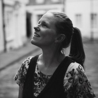 Photo of Kate Arthurs - a smiling woman with tied back light coloured hair, standing in a street, is looking up at her surroundings