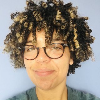 A person with curly, multi-tonal hair poses against a soft blue background, wearing a green shirt.