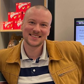 A person in a brown jacket stands casually, with Maltesers boxes visible in the background and a screen displaying event information nearby.