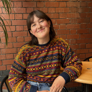 Person in a colorful patterned sweater and light blue jeans sitting on a black chair at a wooden table, with a brick wall background.