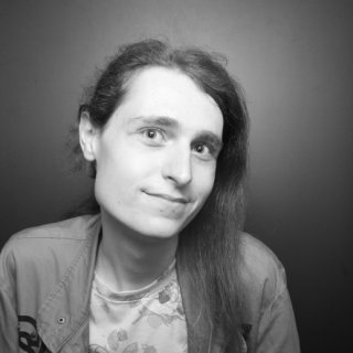 A black and white portrait of a person with long hair looking at the camera with a slight smile. They are wearing a patterned t-shirt under an open jacket.
