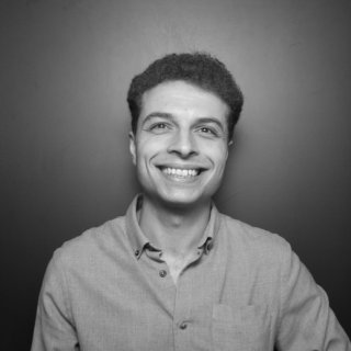 A young man with short, dark, curly hair. He is looking directly at the camera and smiling broadly, showing his teeth. He is wearing a button-down shirt and is set against a plain, dark background