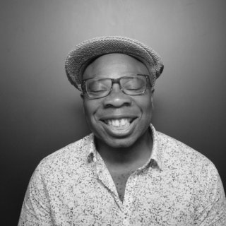 A black and white portrait of a man wearing a cap and glasses, smiling broadly with his eyes closed.