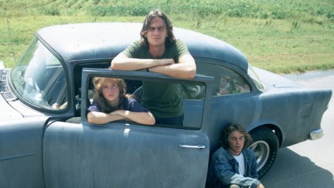 Film still of three people with a parked car, one standing, one in the fron seat and one lying on the road leaning against the car