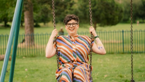 A woman with short hair and glasses wearing a multi coloured, stripey outfit sits on a swing smiling, with trees and grass in background.
