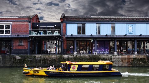 Exterior shot of Watershed building with a cloudy sky and a blue and yellow Bristol Ferry Boat passing on the water.  