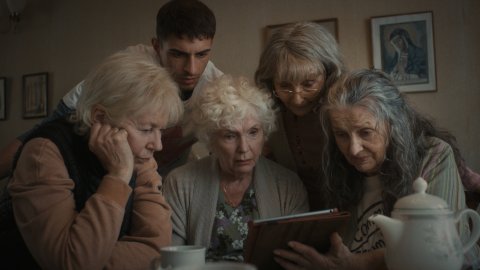 A group of four older women and 30-something man crowd around a tablet
