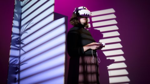 A woman stands with a VR headset on and controllers in here hand, the background is purple and pink with various shadows.